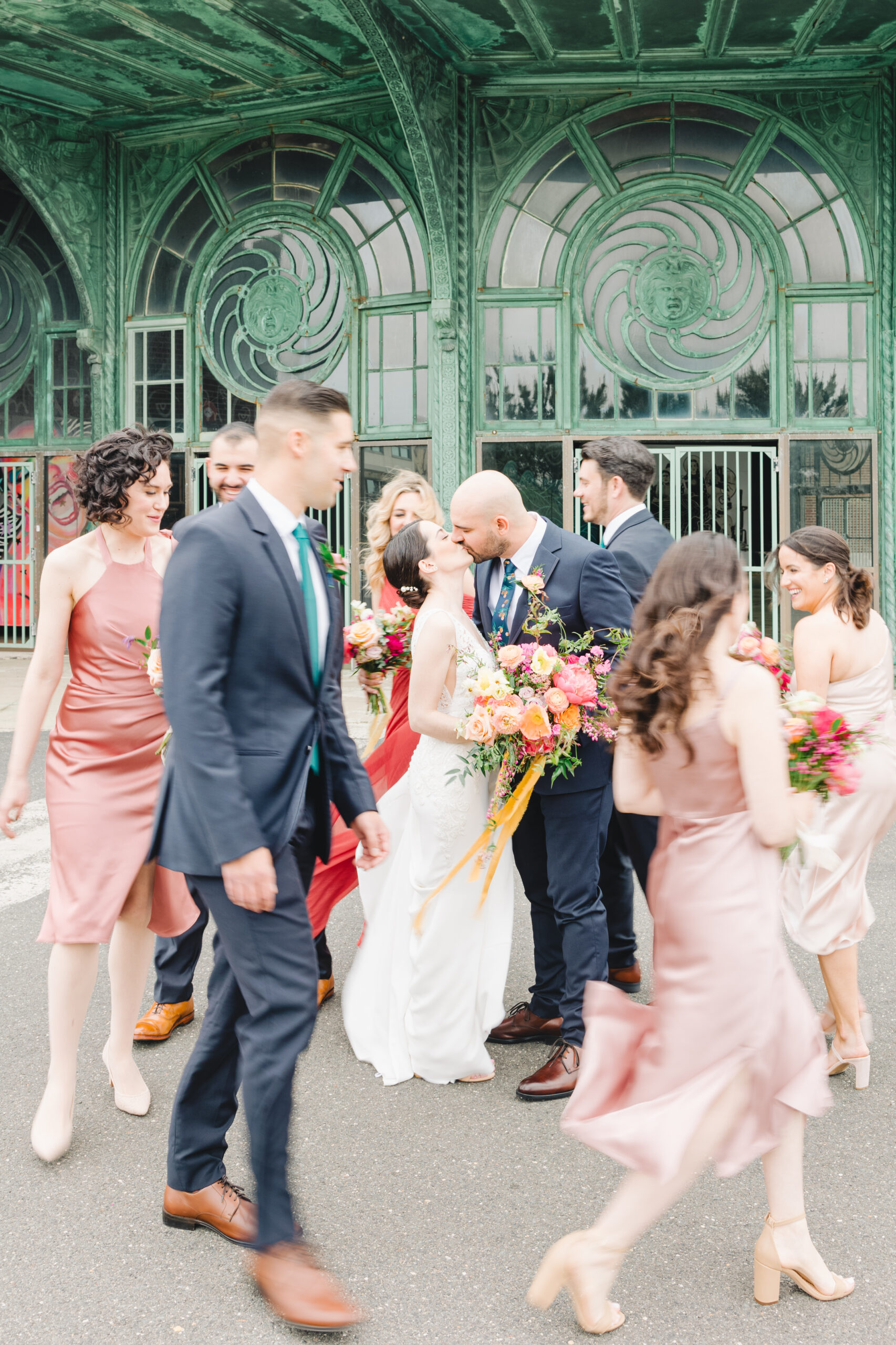 The bride and groom kiss as their wedding party runs by outside the old carousel in Absury Park ahead of their Porta Asbury Park wedding.