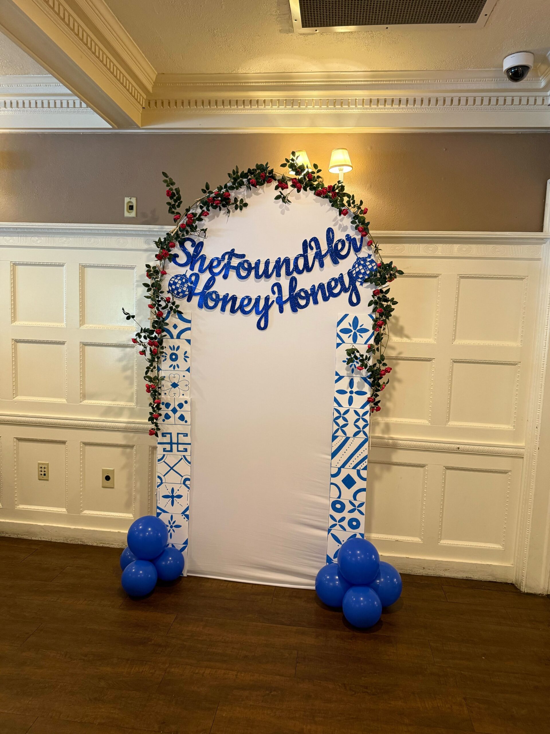 Hand-painted Greek tile backdrop with pink flower garland and "She Found Her Honey Honey" sparkly blue sign at a Mamma Mia bridal shower

