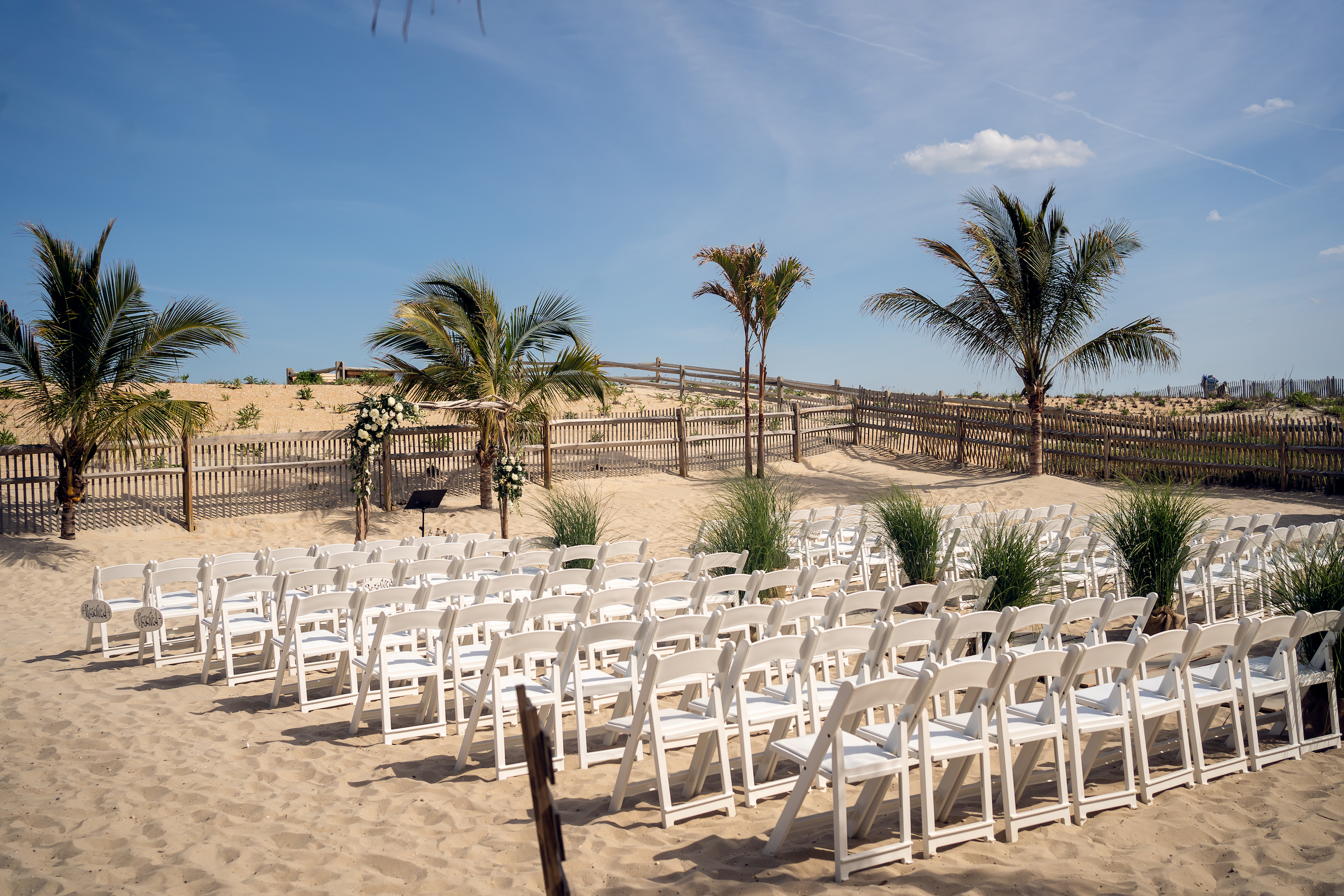 The outdoor ceremony space at Sea Shell Resort and Beach Club features white chairs on the sand with palm trees.