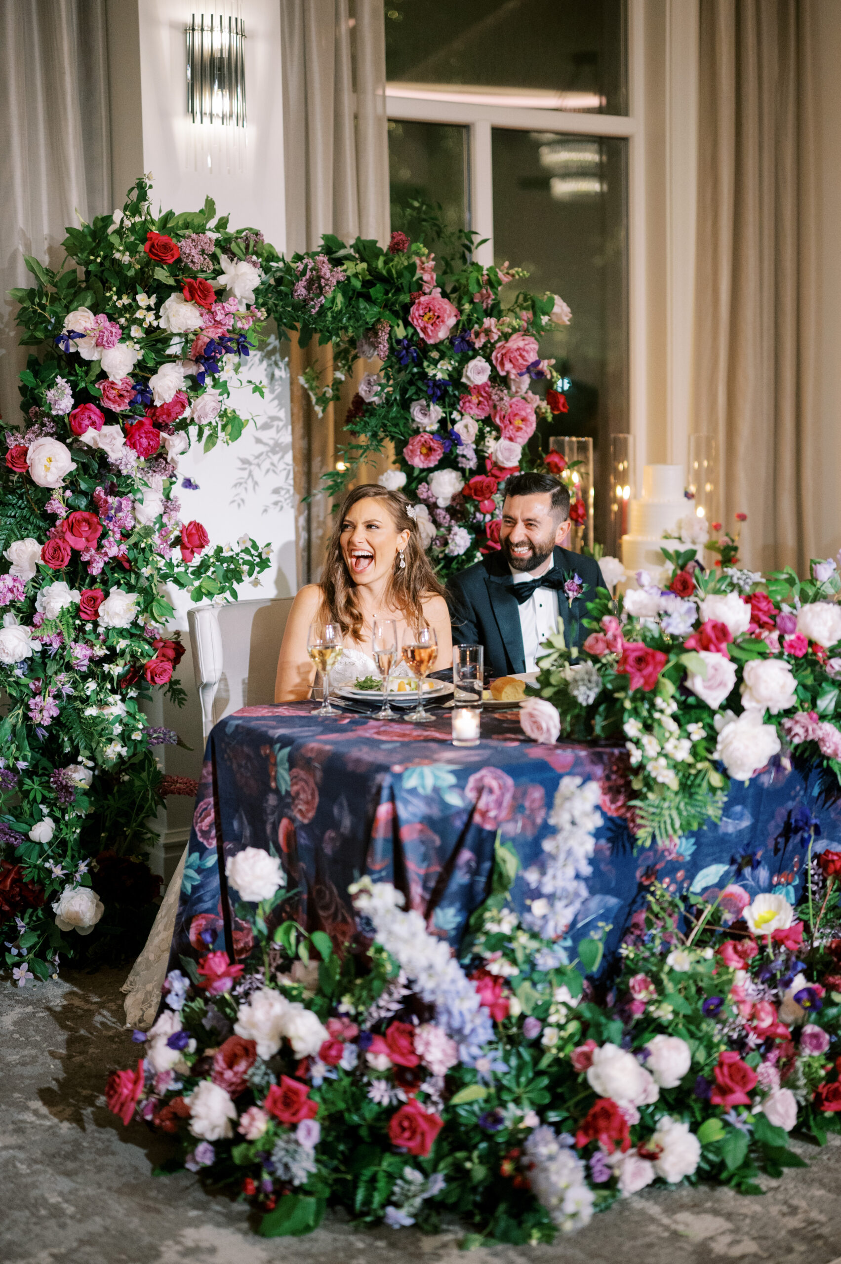 The bride and groom sit at the sweetheart table at their Chateau Grande Hotel wedding which features a purple tablecloth and an abundance of pink and purple flowers on the table and behind the couple.