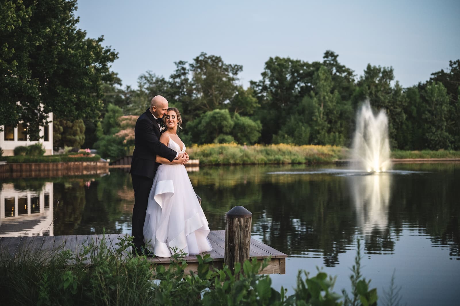 The bride and groom embrace each other on a dock overlooking a lake with a fountain in the background.