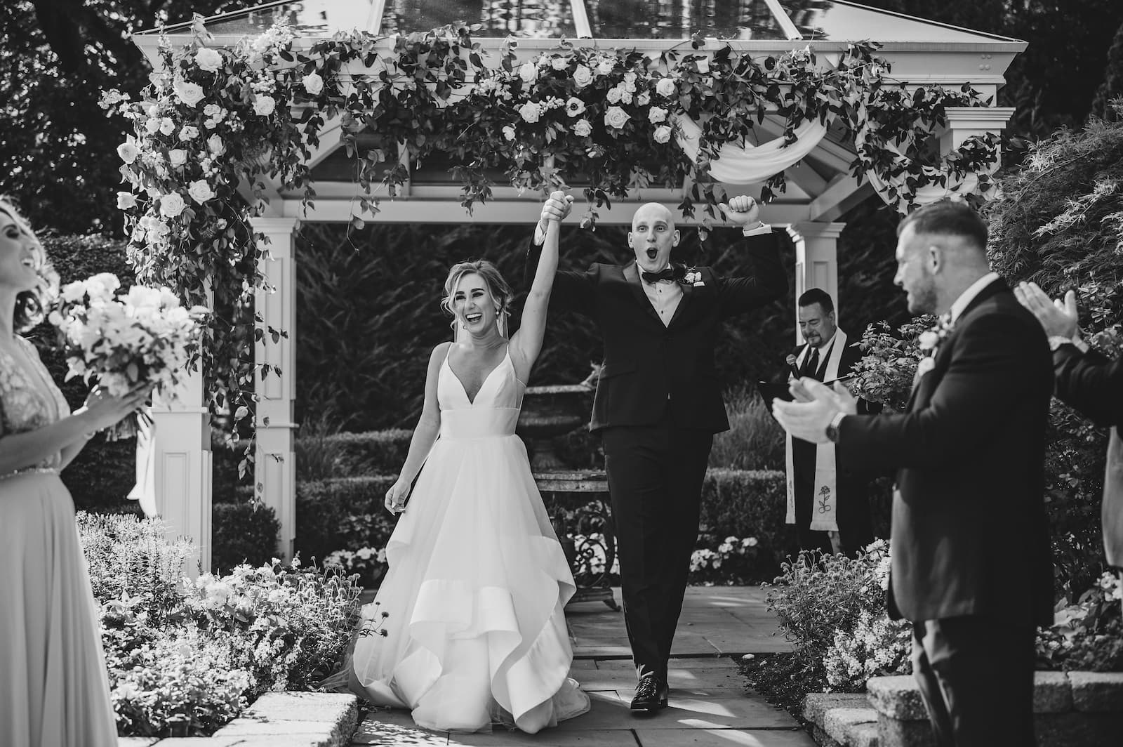 A black and white photo of a bride and groom holding hands and raising them in the air right after getting married with a floral archway in the background.