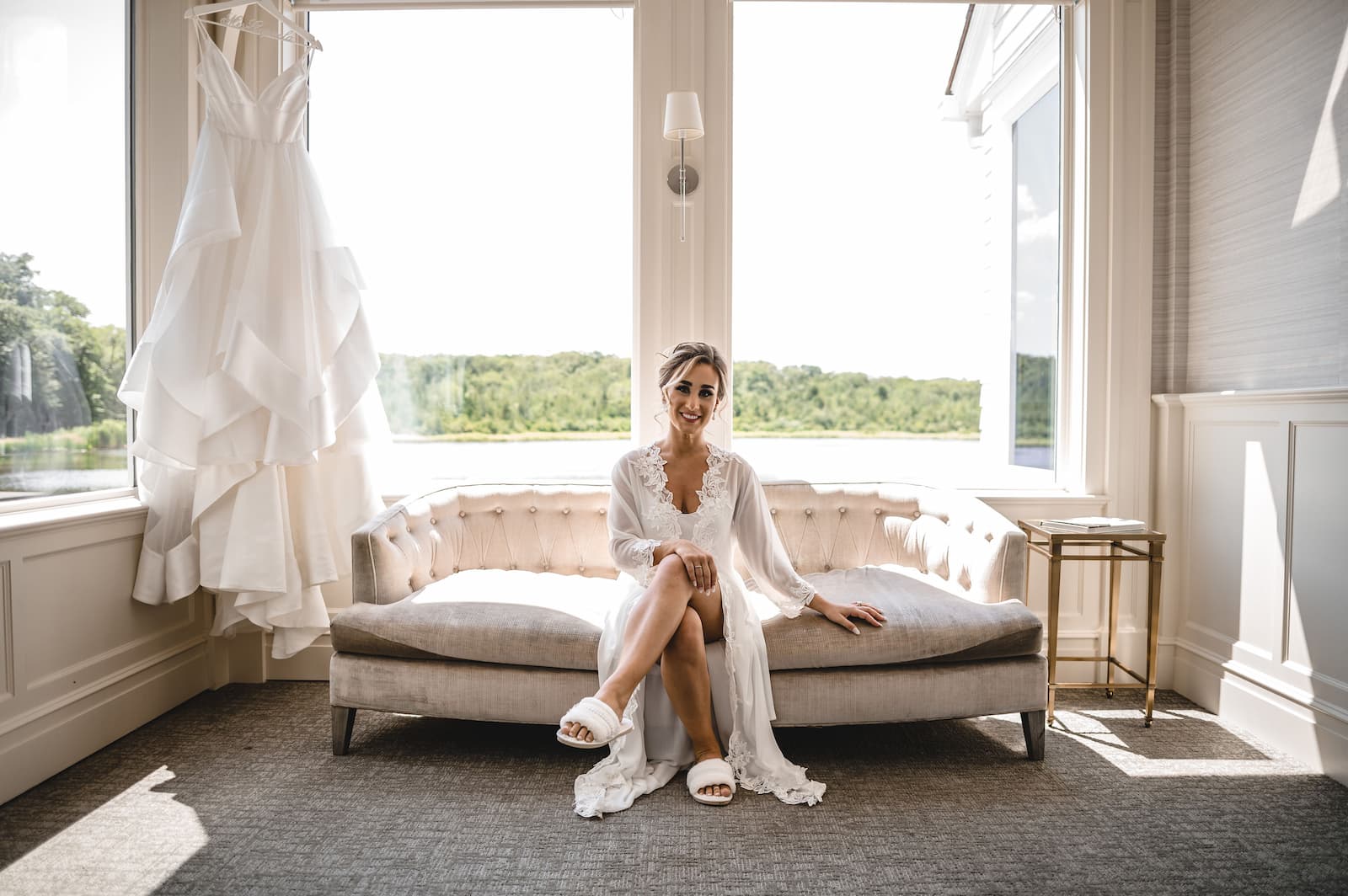 The bride sits on a couch in her getting ready robe with her dress hung up in the background and two big windows overlooking at lake.