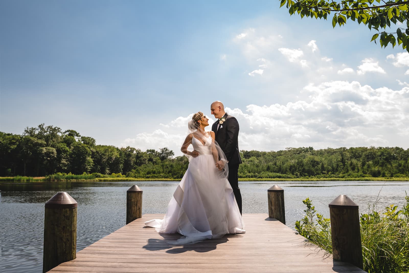 The bride and groom look at each other on a dock overlooking a lake with a blue sky.