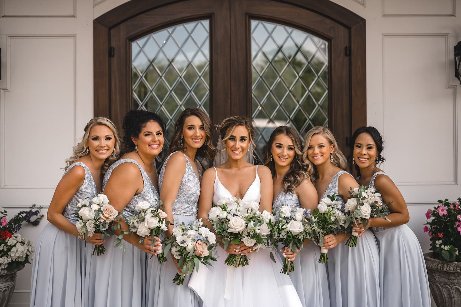 The bride is flanked by her bridesmaids with pale blue dress and bouquets in front of brown doors with windows.