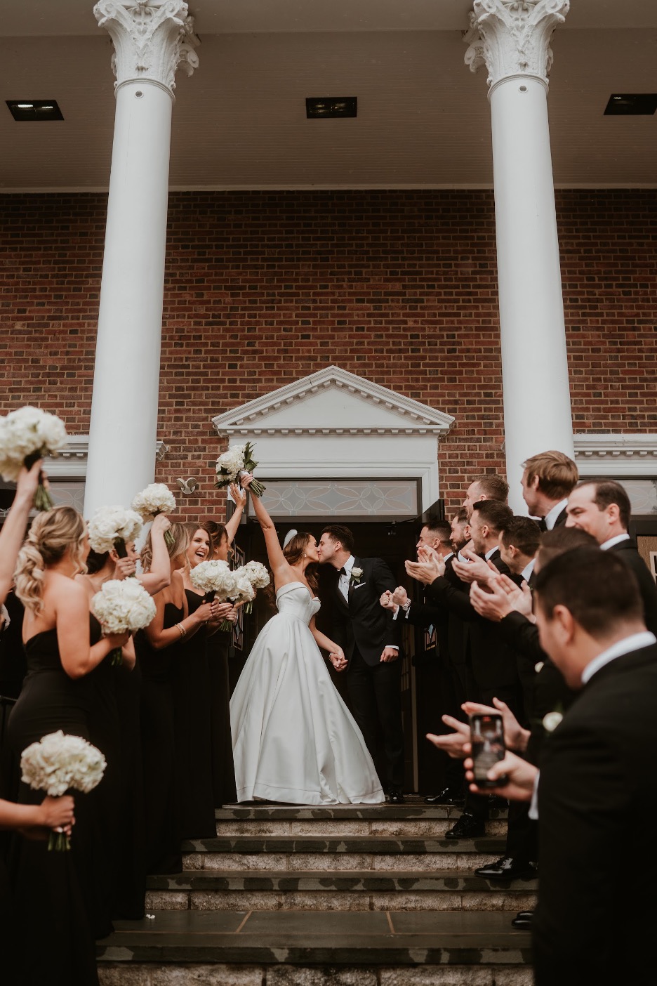 The bride raises her hand holing the bouquet as she kisses the groom outside of their ceremony with a brick wall and columns behind them and the wedding party gathered on the steps cheering.