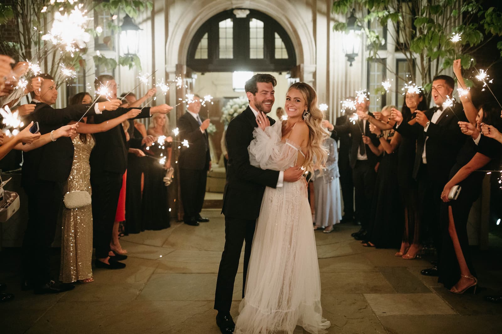 The bride and groom pose during their sparkly send-of outside the venue with guest lining the pathway holding the sparklers.