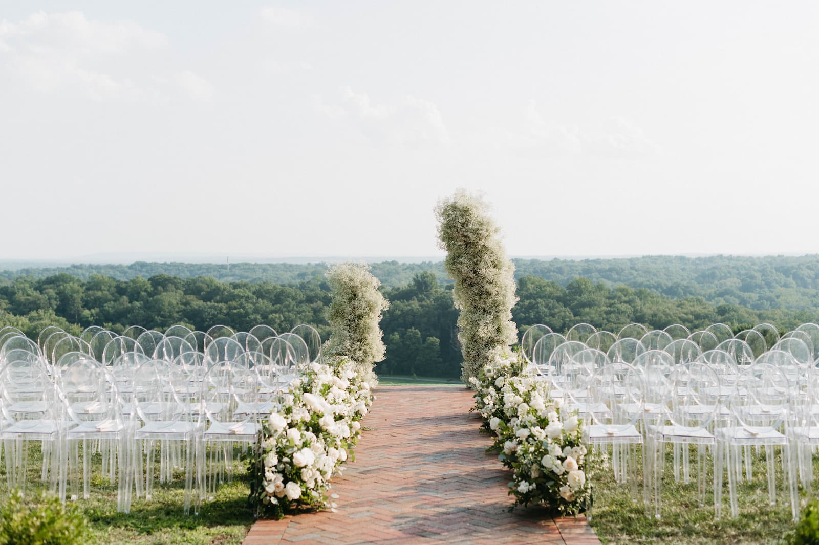 Rows of clear chairs set up for the ceremony outside overlooking rolling hills and a two piece floral archway in the front with white and green flowers.
