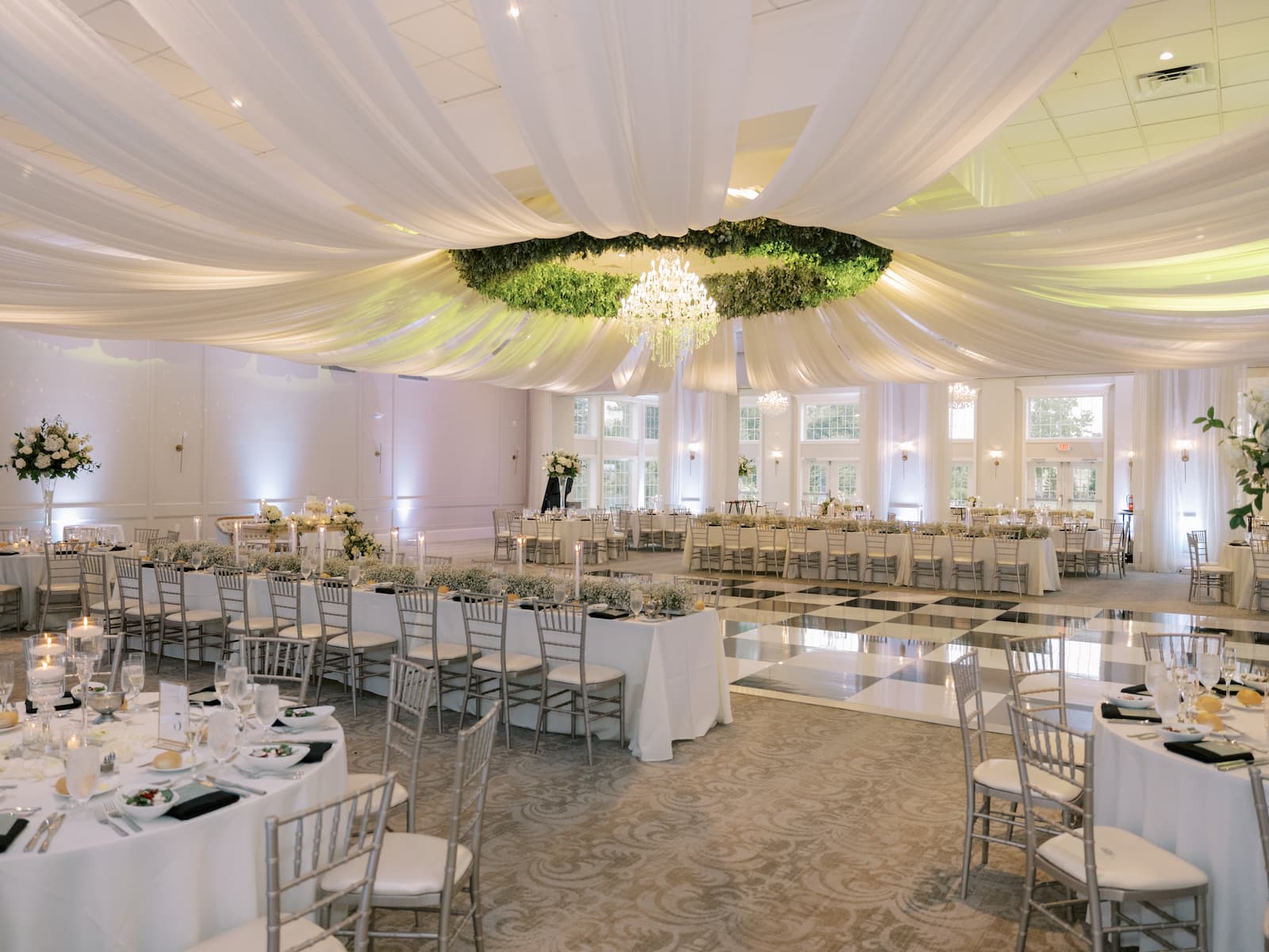 The neutral colored ballroom features a large chandelier surrounded by greenery and large draping clothes cascading out from the chandelier.