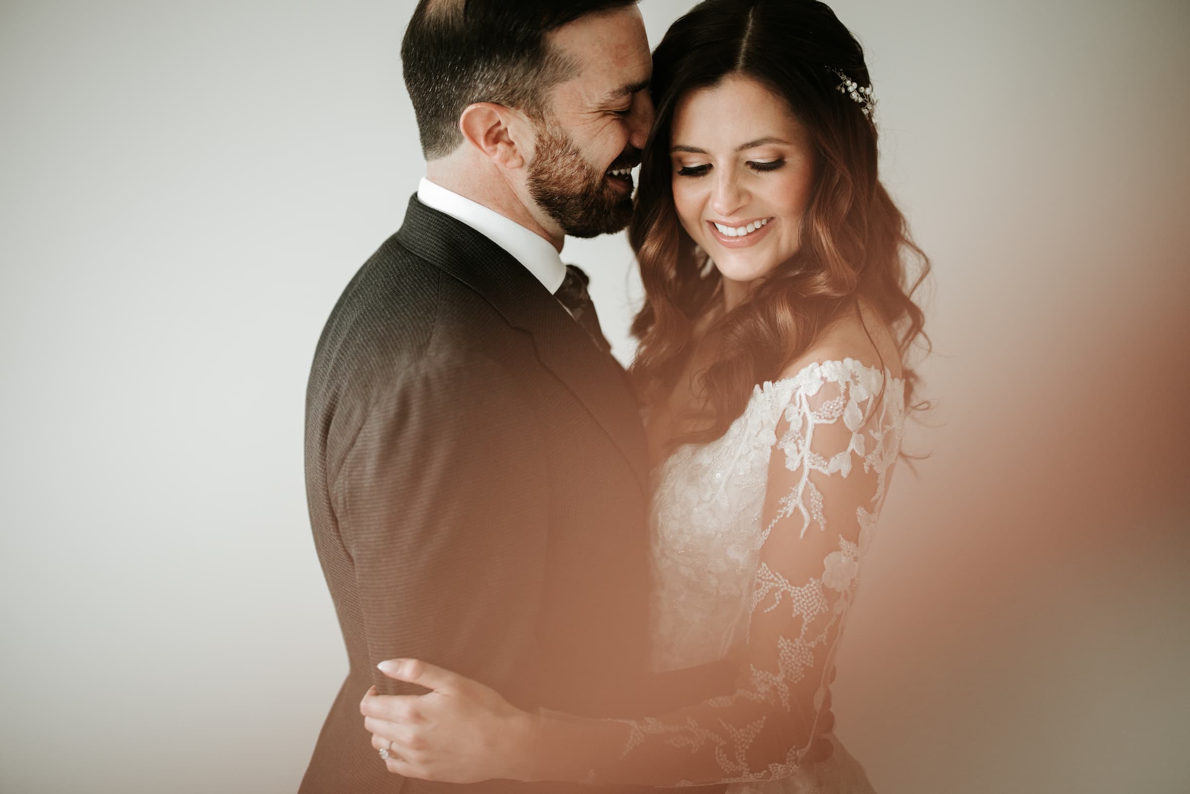 The bride and groom embrace while facing each other. The bride is looking down and smiling towards the camera. The groom nuzzles his nose into her temple and smiles.