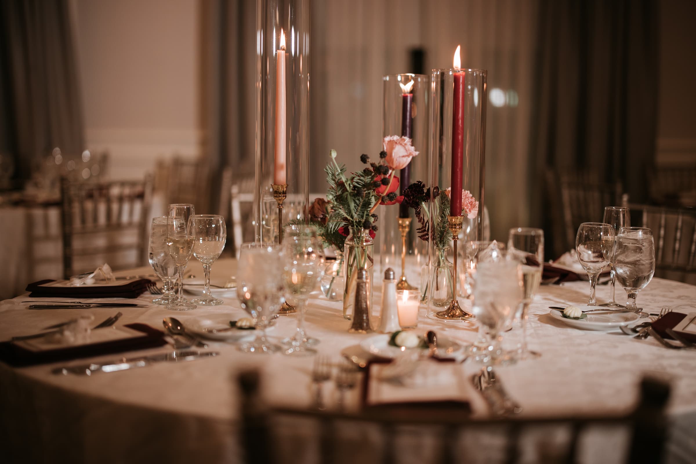 A closeup of a tablescape with tall red candles, flowers of various shades of red in a vase and glassware.