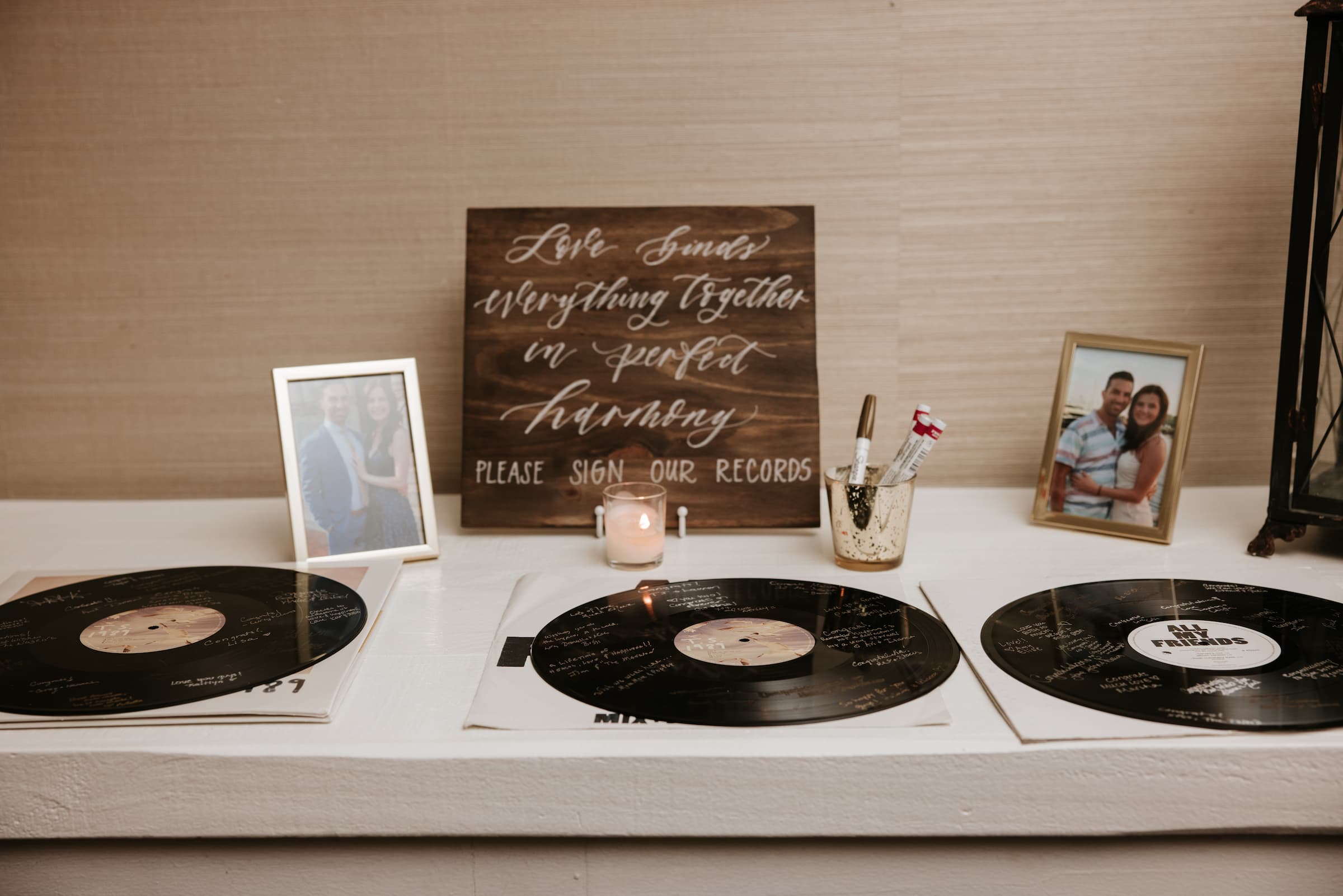 Three records are laying flat on the table with a wooden sign behind that reads "Love binds everything together in perfect harmony. Please sign our records." There is one framed photo of the couple on each side of the sign, a different take on a guest book.