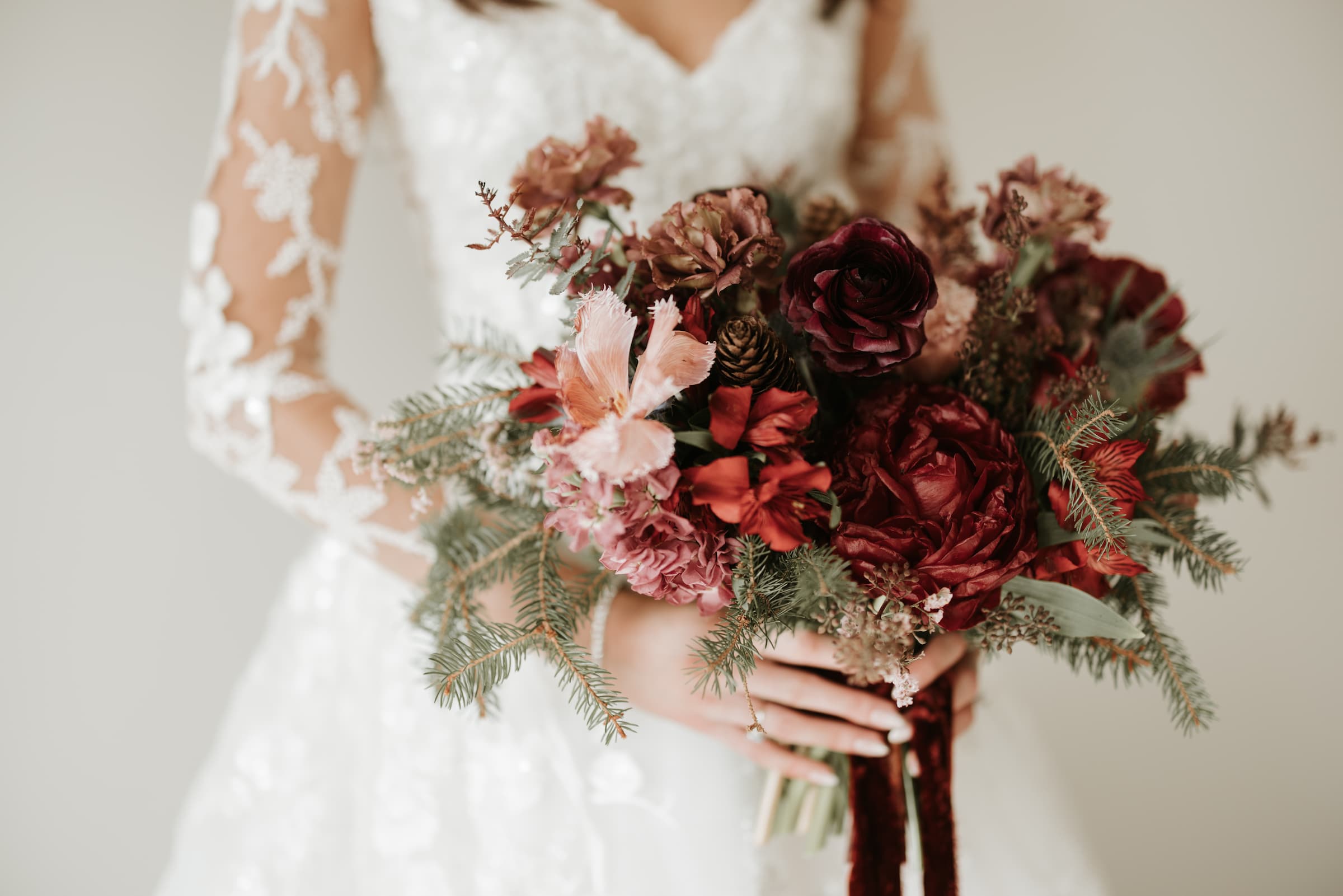 A closeup of a burgundy, red and pink bouquet in focus held by a bride in a white gown with lace sleeves.