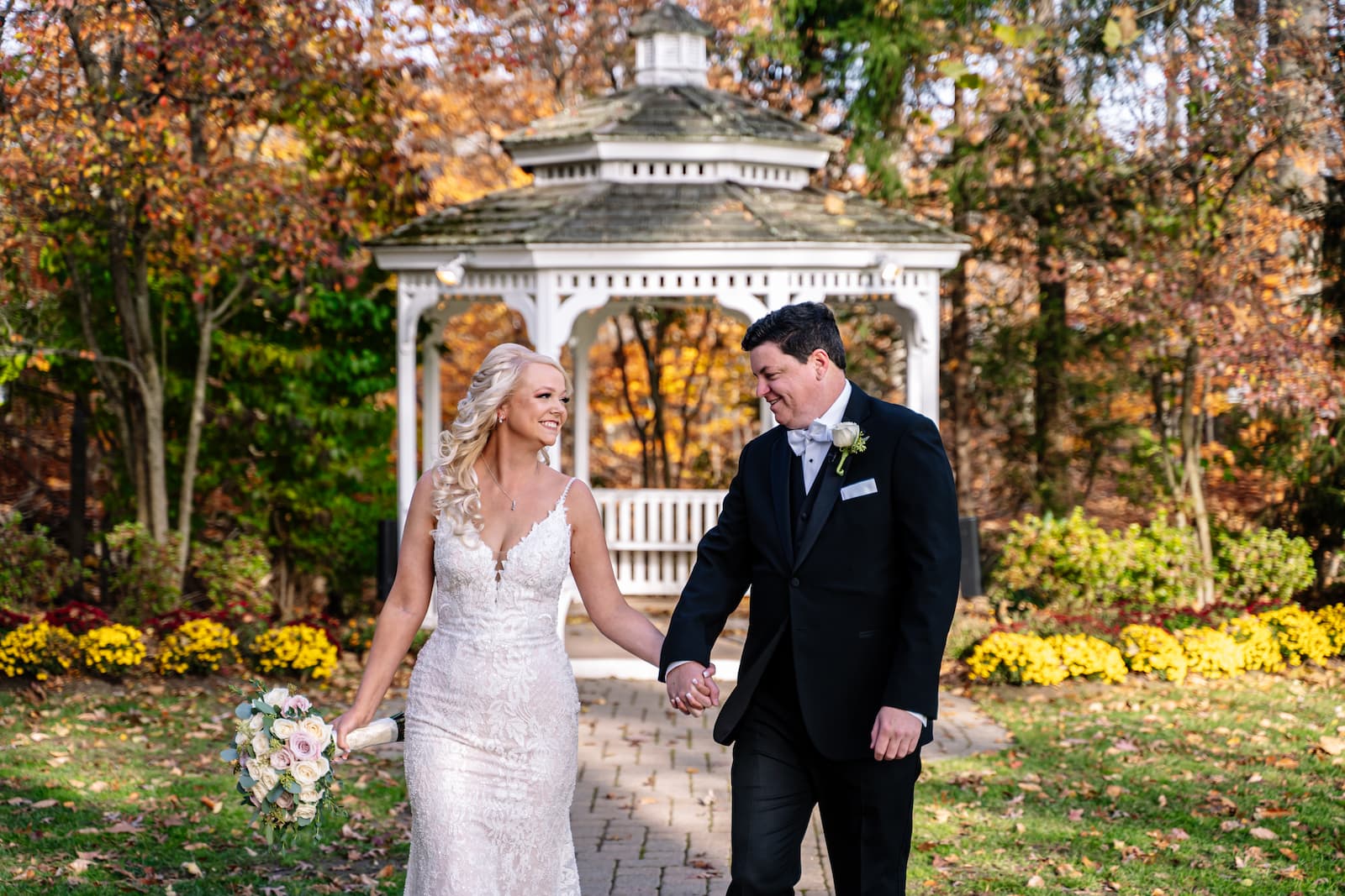 The couple hold hands and smile as they walk down a stone path with a white gazebo in the background and orange and green trees.