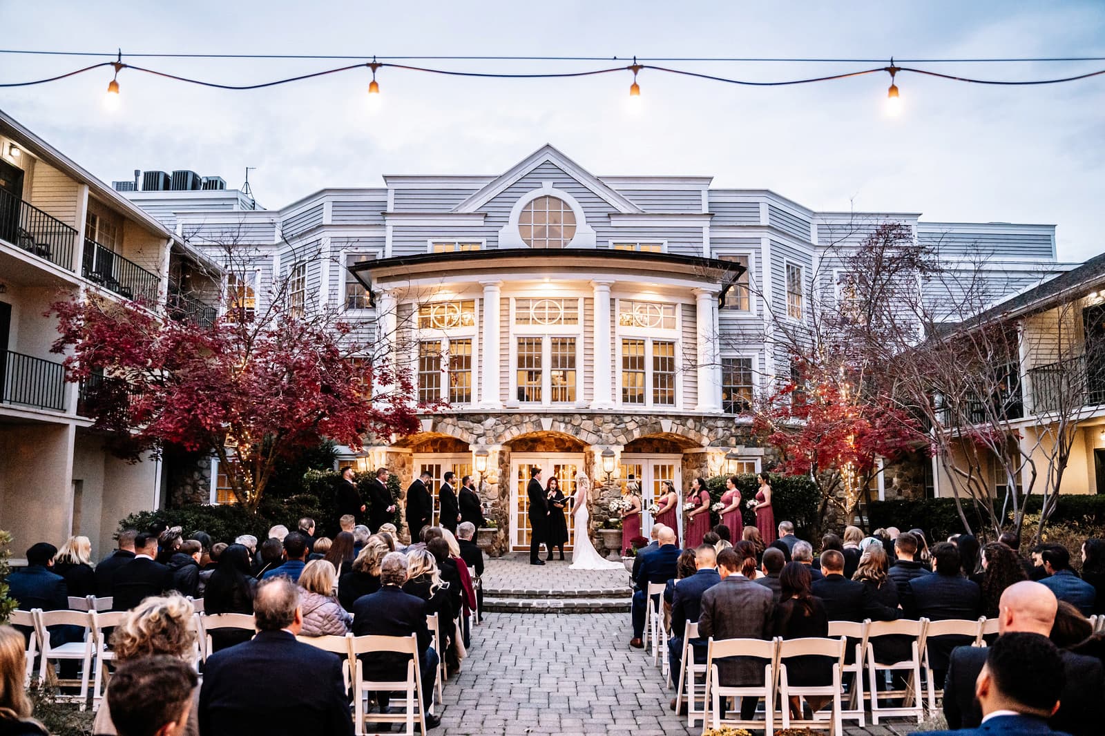 A pulled back shot of the outdoor ceremony. The couples stands two steps up from the guests on a stone path. The stone archway of the building is behind them. Surrounding the ceremony area is the venue's hotel rooms. Guests sit in beige folding chairs.