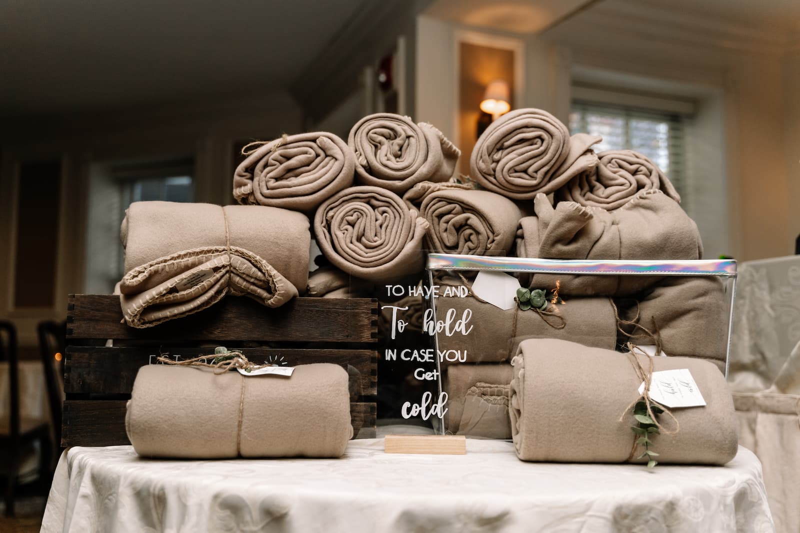 A pile of rolled and tied brown blankets are stacked on a table behind a sign that read "To Have and To Hold In Case you Get Cold."