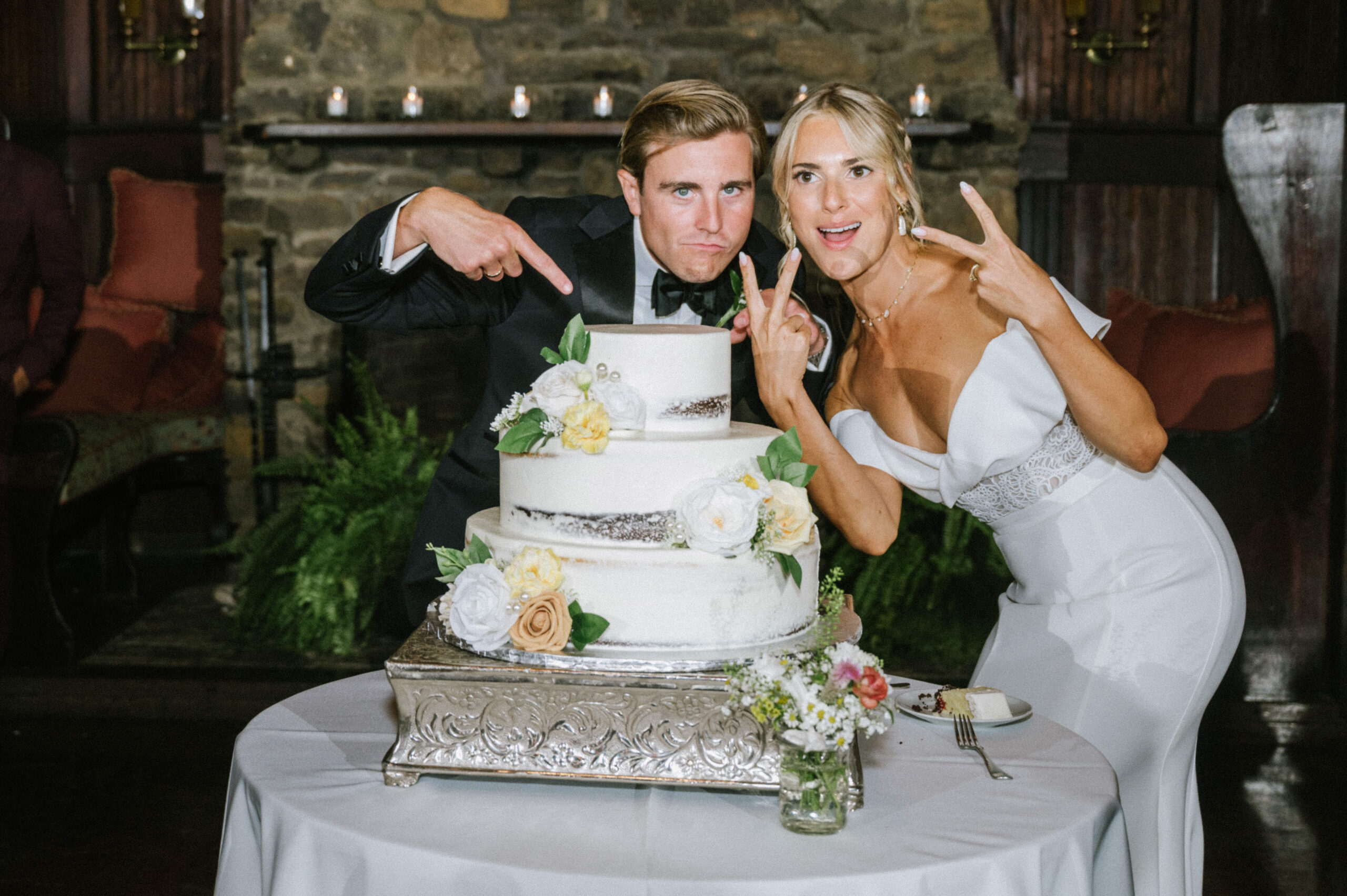 The bride and groom make silly faces and pose with their white wedding cake with yellow, white and green florals at Water Witch Club.