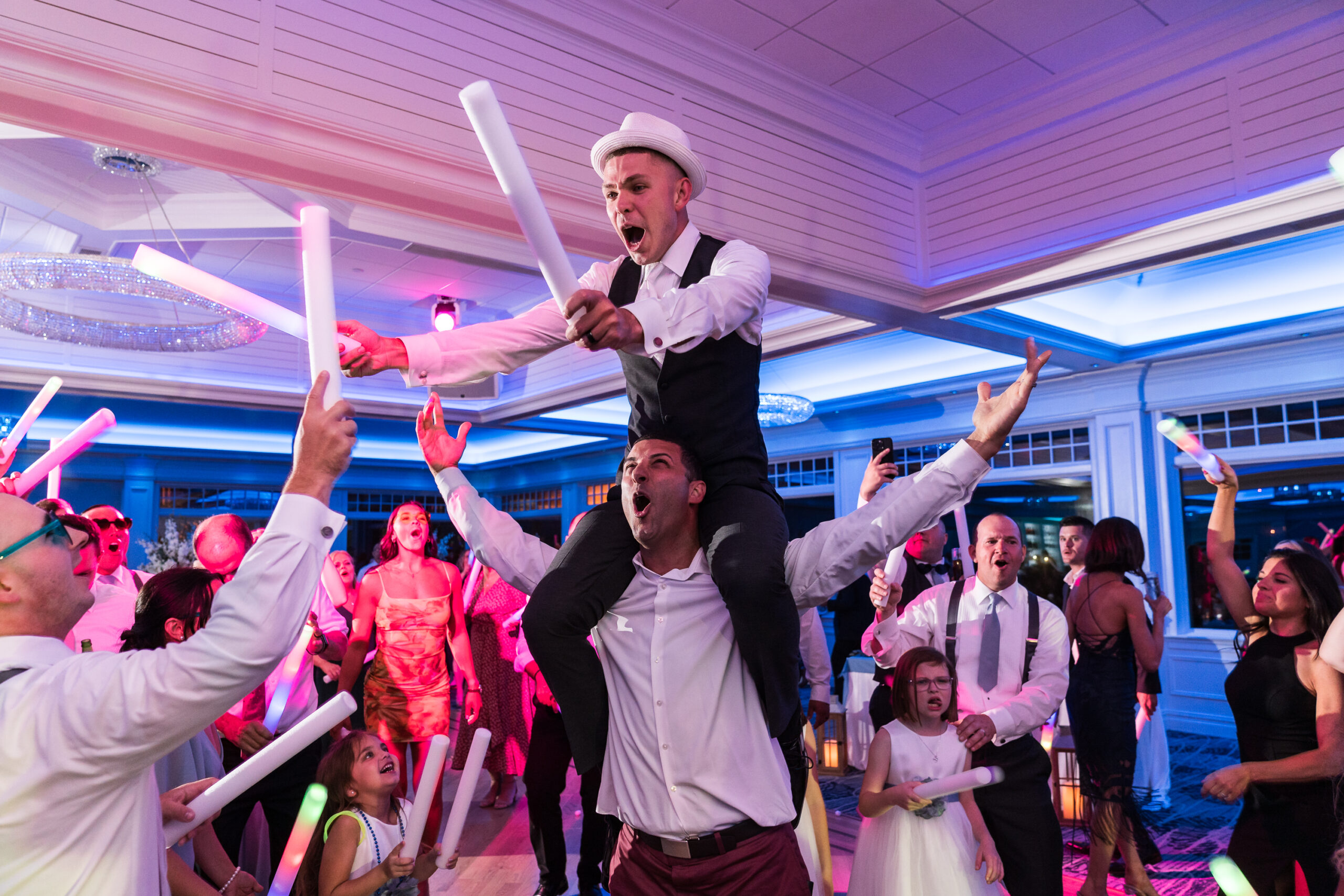 The groom dons a hat as he sits atop a friends shoulders on the dance floor at his Crystal Point Yacht Club wedding.