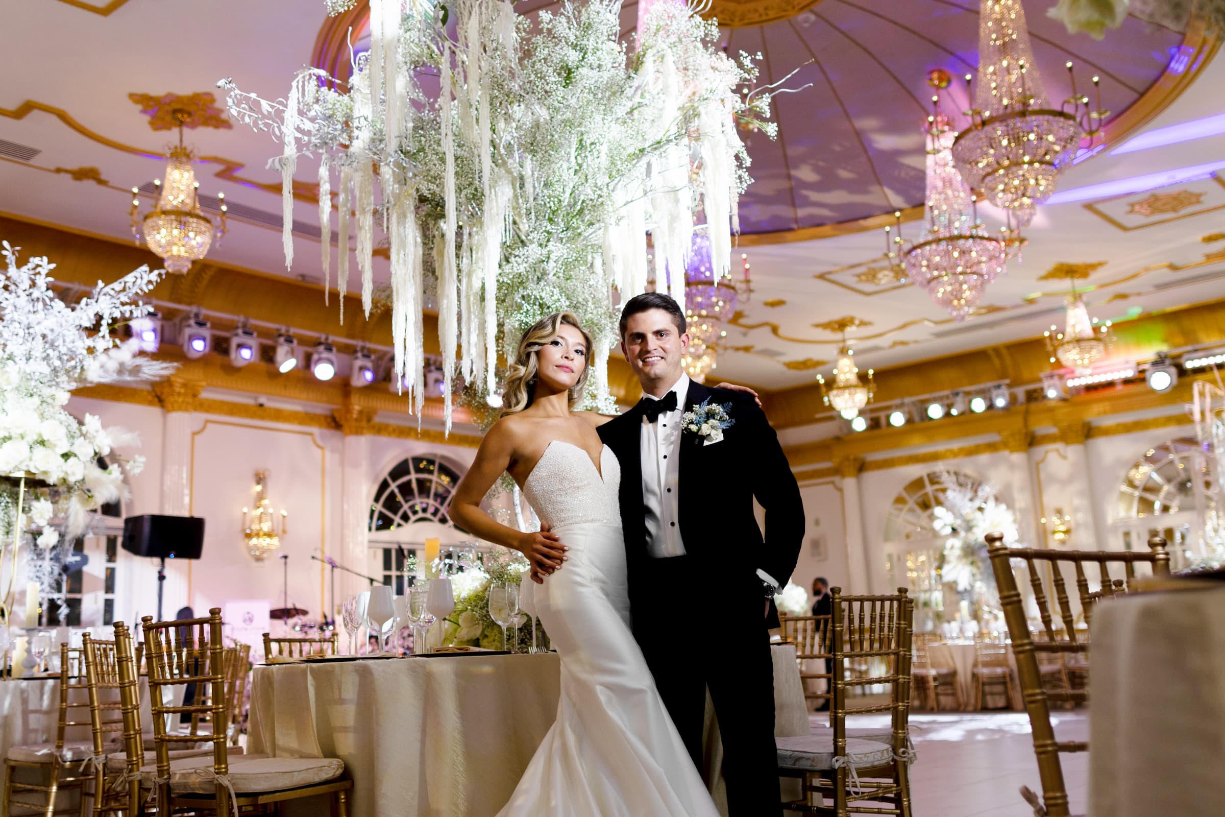 In front of a very tall, waterfall-like centerpiece that resembles a tree with long white decorations hanging off of it, the bride and groom pose with their around each other in an otherwise empty ballroom.