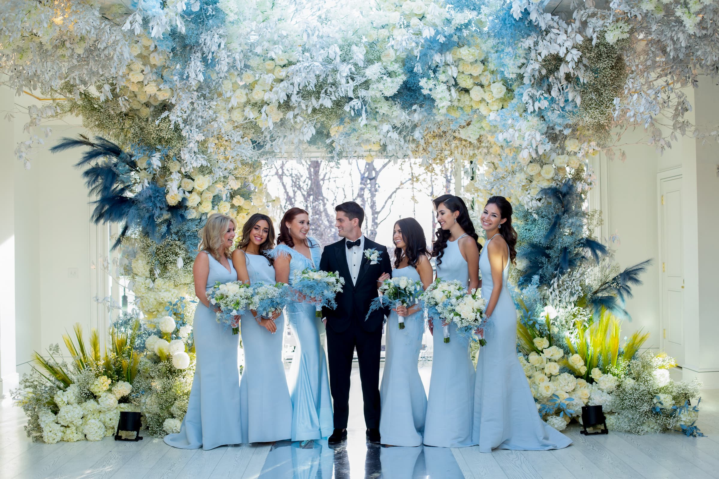 The groom in a black tux is flanked by three bridesmaids on each side wearing pale blues gowns. The backdrop is an elaborate blue and green floral archway.