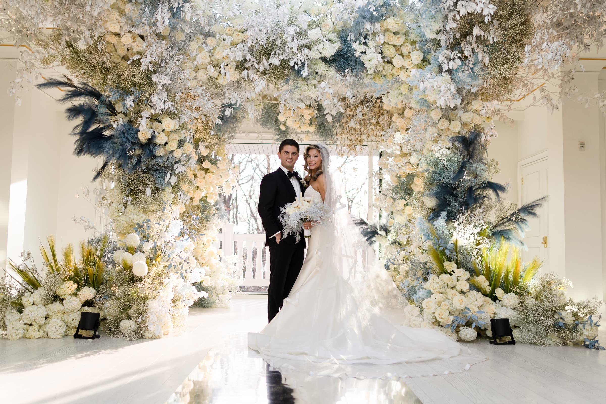 A portrait of the bride and groom in front of elaborate blue, white and yellow florals, a glass ceiling and a mirrored reflective floor.