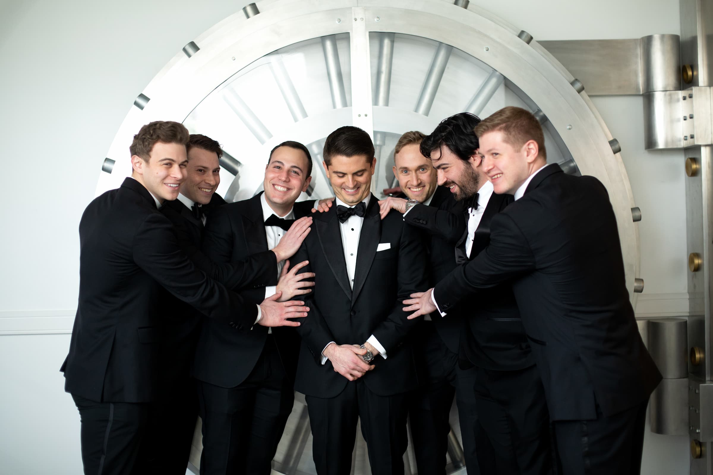 The groomsmen decked out in black tuxes smile and embrace the groom as they stand in front of a vault-like background.