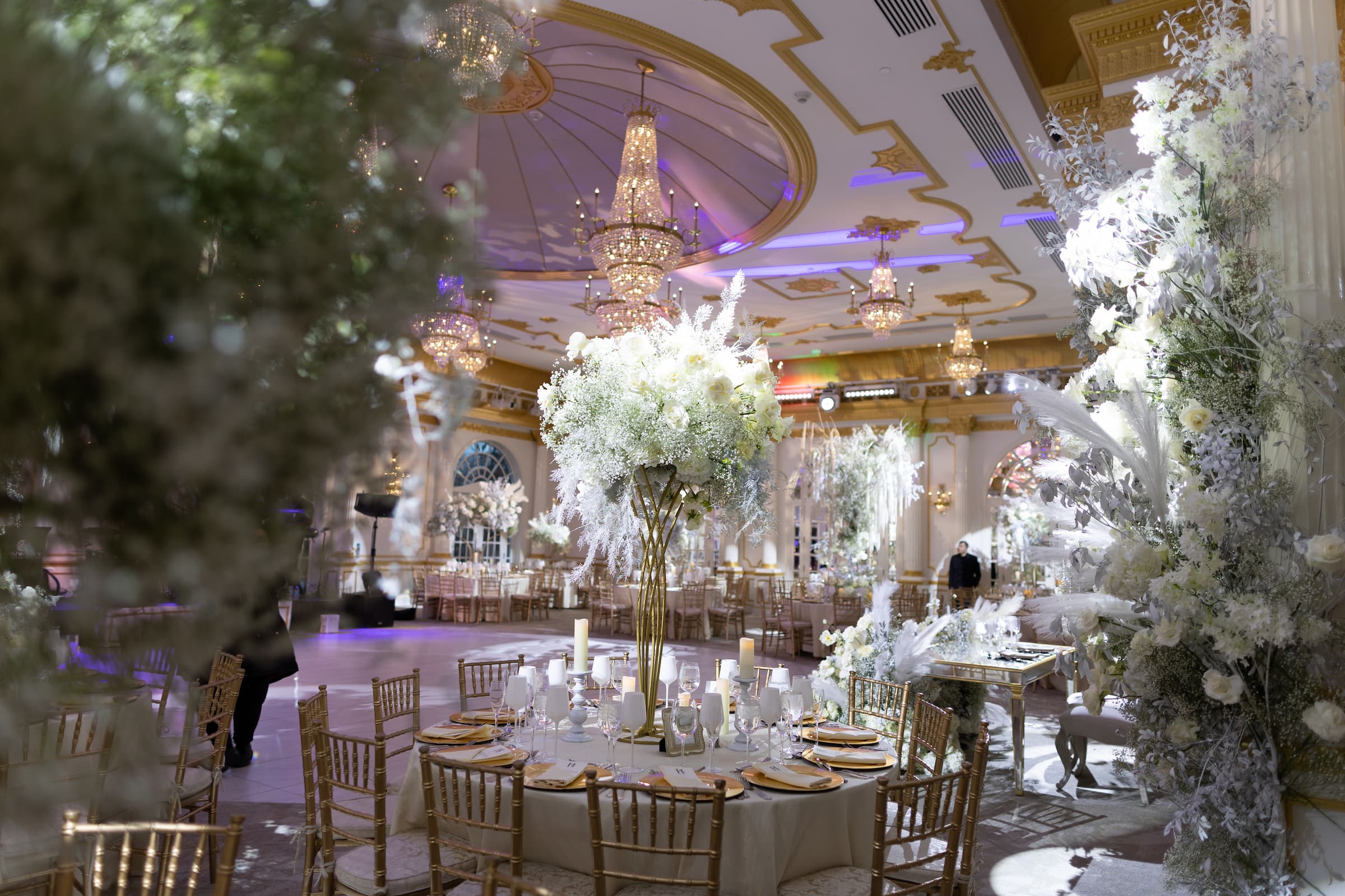 A tall golden centerpiece holds white flowers among a tablescape with candles and glassware.