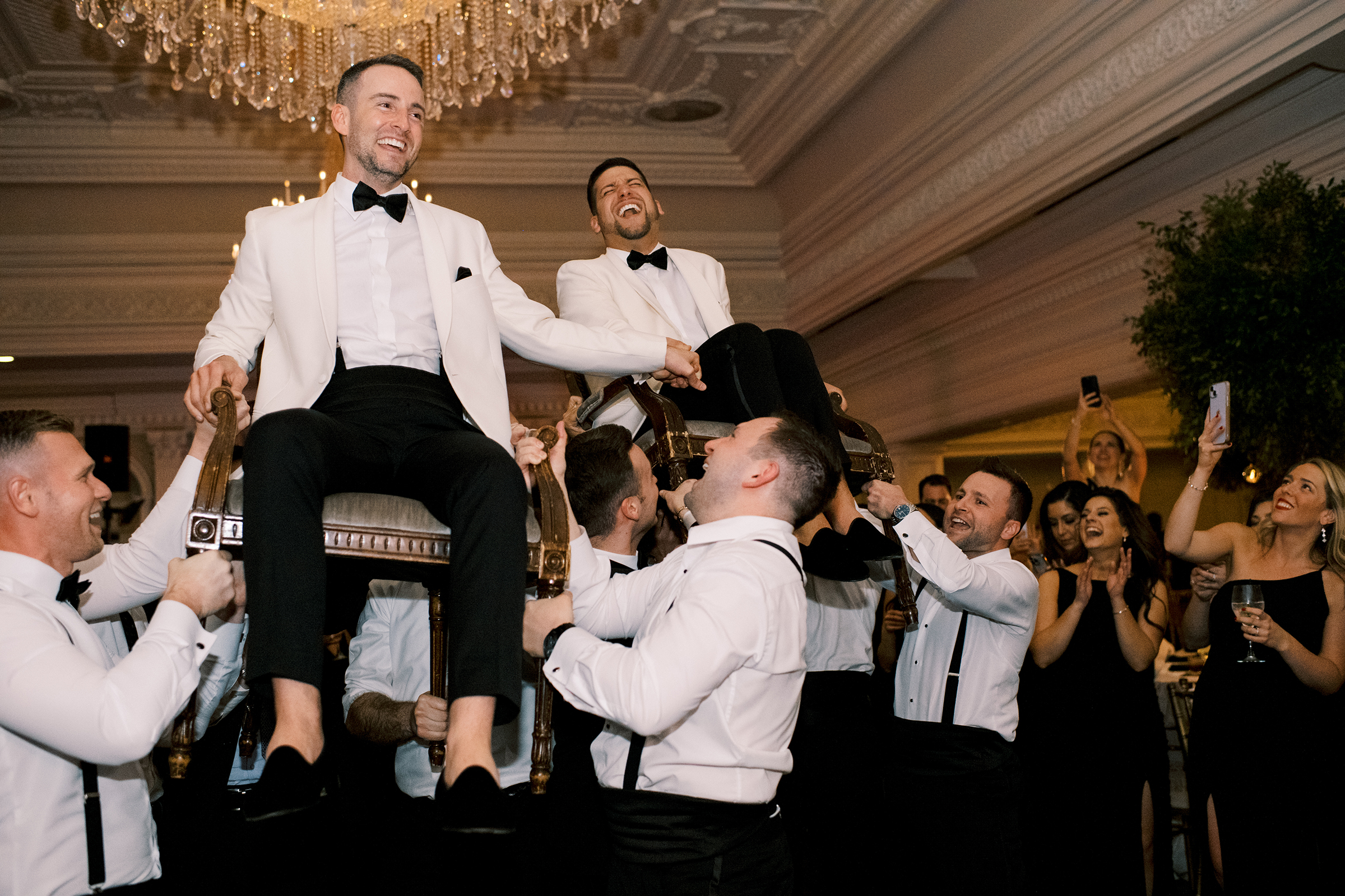 The grooms, in ivory tuxes, laugh as guests hold them up in chairs during the reception at their Park Savoy Estate wedding.