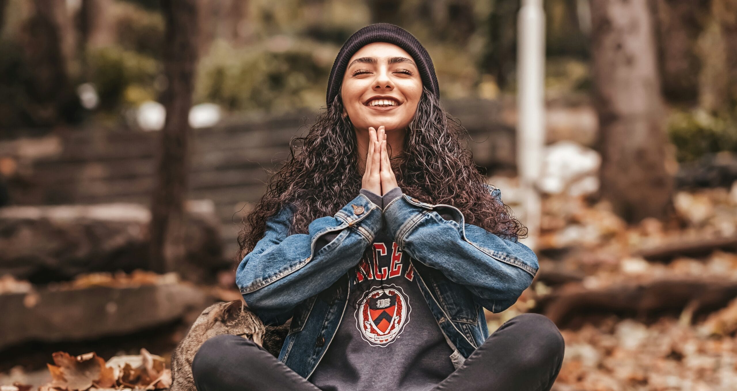 A woman sitting in the forest with her legs crossed and her hands in a prayer position with a smile on her face.