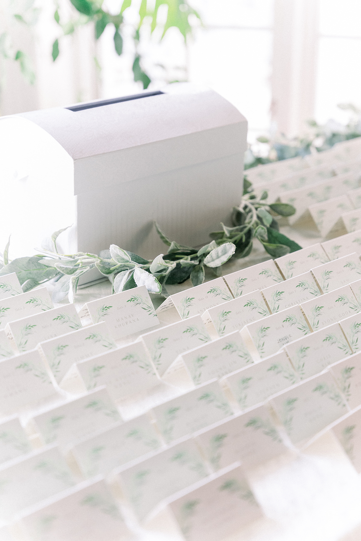 A simple white card box surrounded by greenery with white and green place cards spread out in front at this Madison Hotel wedding.