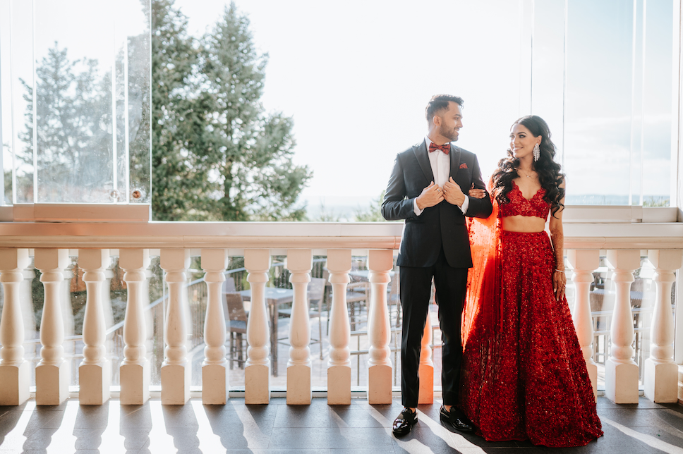 The groom in black and the bride in red stand by a balcony at their Westmount Country Club wedding.